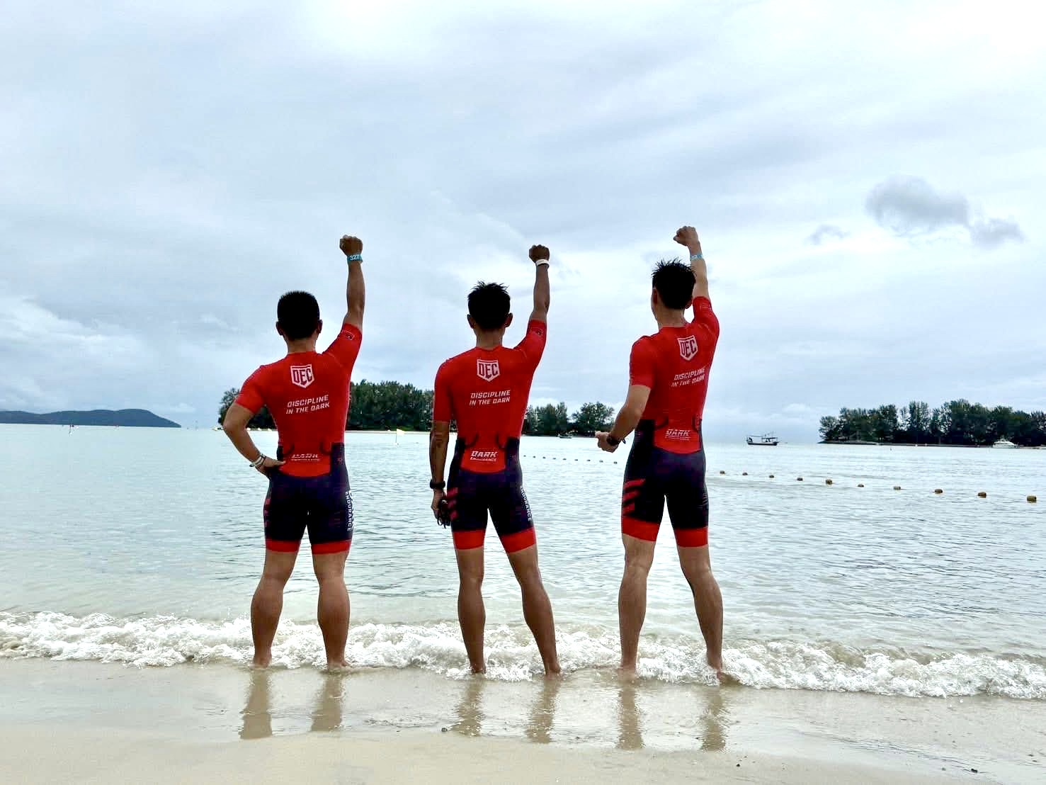 Three DEC athletes in race kit, fists raised on a beach